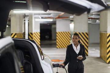 Focus on attractive lady with cell phone standing near electric automobile with charge cable in port. African entrepreneur in business suit preparing battery powered car at parking for trip.