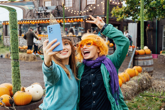 Two laughing female friends in fun glasses and witch hat having fun and making selfie on phone on decorated pumpkin farm. Selecting Thanksgiving and Halloween holidays decor. Autumn fall festive mood