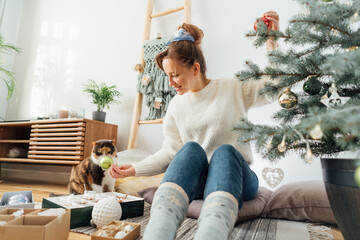 Young woman in cozy sweater decorating potted Christmas tree while her cat is watching her in light...