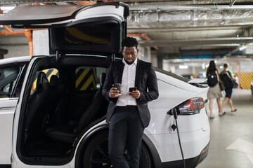 Wide view of basement parking garage where african american man having drink and checking phone calls during EV fill-up. Relaxed adult using coffee break for powering electric motorcar at workplace.