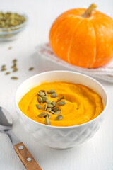 Homemade vegetarian creamy pumpkin soup made of pureed vegetables decorated with peeled dried seeds served in bowl with spoon on white wooden table with orange color squash is a Thanksgiving dish