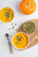 Top view of homemade vegetarian pumpkin soup made of pureed vegetables decorated with peeled dried seeds served in bowls with spoon on white wooden table with ingredients is a Thanksgiving dish