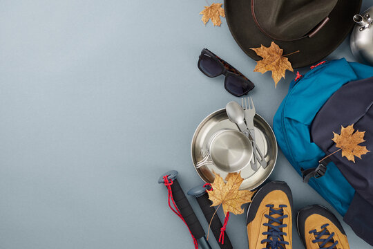 Packing for a hiking trip in the fall. Top view flat lay of different camping equipment, hat, sunglasses, fallen leaves on blue background with commercial placeholder