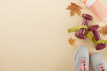 Autumn style and fitness harmony. Top view photo of stylish sneakers, measuring tape, dumbbells, plastic water bottle, fallen maple leaves on pastel brown background with promo spot © Goncharuk film