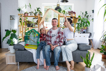 traditional family with a child and a parrot pet in a cage are enjoying a new home, sitting on the sofa and holding the roof. Mortgage, insurance, buying and moving into your own home