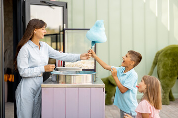 Little girl and boy waiting for a sweet cotton candy to be made by female seller at outdoor shop of...