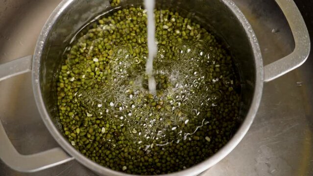 Cooking mung beans by pouring water into a steel pan with green grains for washing and soaking close up