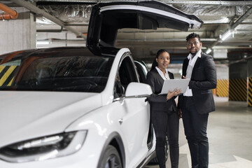 African american business partners in formal clothes viewing information on digital screen near e-car. Pretty woman and handsome man planning presentation with help of laptop after work in office.