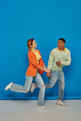 Naklejka premium happy multicultural couple in casual attire holding hands and running on blue background, smile