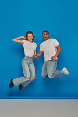 Fototapeta na wymiar positive multicultural couple holding hands and jumping on blue background, full length view