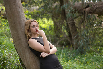 Young woman, blonde and beautiful, with a black dress, leaning on the trunk of a tree, in the...