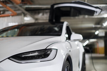 Focus on headlight of modern white electric vehicle being parked with open wing door in underground garage. Battery-powered automobile being supplied with renewable energy via charging cable.
