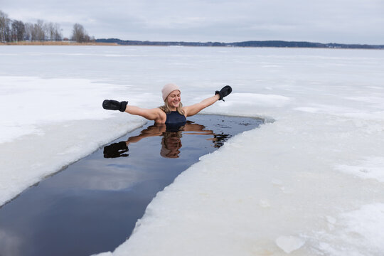 Winter swimming. Woman in frozen lake ice hole. Swimmers wellness in icy water. How to swim in cold water. Beautiful young female in zen meditation. Gray hat and gloves swimming clothes. Nature lake