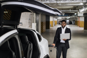 Smiling middle-aged man in formal suit plugging in EV charging cord while carrying computer in hand...