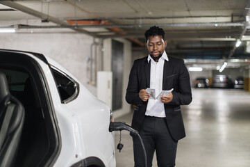 African male adult in business suit thumbing through cash while supplying battery-driven vehicle in indoor charge point. Handsome parking customer reminding of recent spending for EV charging.