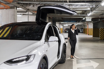 Charming female in business suit speaking on cell phone while refueling e-car via charger gun in underground parking. African american lady exchanging experiences of EV usage for short trips.