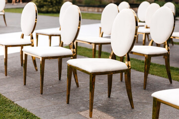 Luxury white-golden chairs on wedding ceremony outdoors. Festive decorations. Empty rows armchairs for guests. Outgoing wedding ceremony in the open air. Decor Studio.
