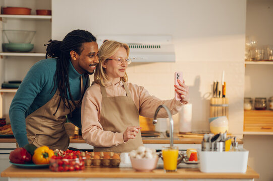 A young multicultural couple is taking selfies while cooking a meal at home