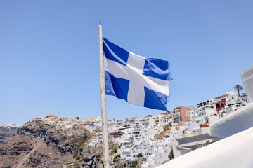 Fira, Greece - July 20, 2023: The Greek flag flying amongst the cliffside buildings and walkways in...