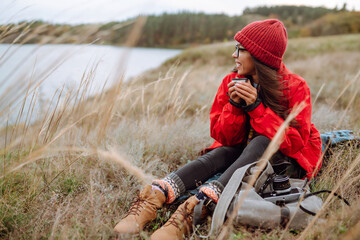 Beautiful woman in a red jacket and a red hat sits in the autumn forest and drinks a hot drink from...