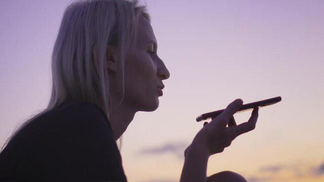 Happy young woman dictating message with street voice recognition on smartphone. sends audio message using your smartphone. Sunset and sky against background of sea on beach.