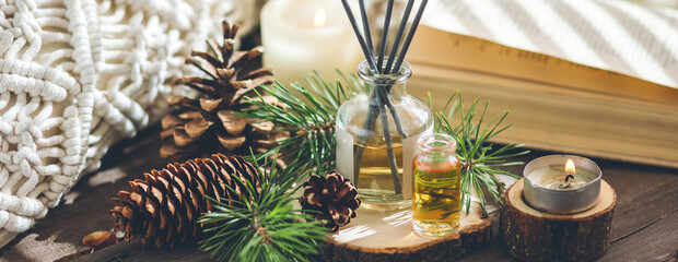 Natural aromatic scented reed diffuser air freshener bottle on wooden table with pine cone, burning candles. Christmas inspiration and mood, cozy home atmosphere, relax. Aromatherapy and decor banner - obrazy, fototapety, plakaty