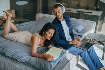 Young bearded American man in headphones works home with laptop wearing headphones looks at wife diary at bedroom. Brunette woman planning agenda laying on bed. Couple remote working home.