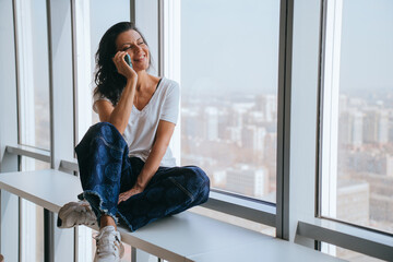 Eyes closed hispanic girl in casual sitting on windowsill talks by phone smiles enjoying pleasant conversation with boyfriend. American young woman at hotel looks at view on city during phone call.