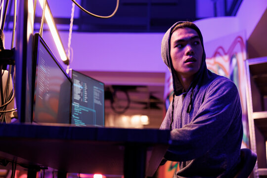 Nervous Young Man Looking Over Shoulder While Breaking Law And Hacking Database In Abandoned Warehouse. Scared Asian Hacker In Hood Afraid Of Police While Coding Illegal Malicious Software