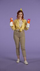 Vertical photo. Woman in casual clothing and rubber gloves holding two bottles of cleansers, smiling looking at the camera.