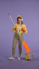 Vertical photo. Woman in casual clothing and rubber gloves in headphones listening music, holding mop, shows thumbs up.