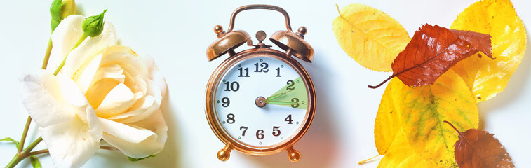 Vintage alarm clock showing the hour between daylight saving time in summer and fall back symbolized by a rose on one side and autumn leaves on the other, panoramic format - 649482242