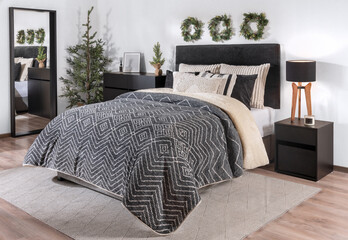 Modern Scandinavian Bedroom Interior with a Padded Fabric Headboard, Warm Black Cover, and Striped cozy Pillows on the Bed, beside a Wooden Nightstand Featuring Christmas Decorations.