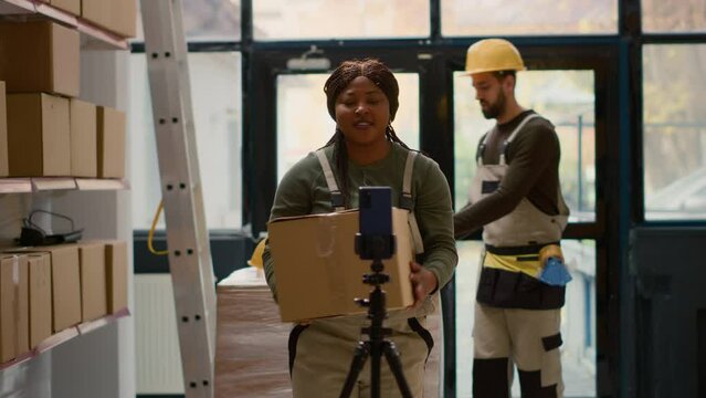 Warehouse supervisor pressing play on cellphone placed on tripod to start recording training video for interns. Quality assurance manager filming tutorial showing trainees how to arrange parcels