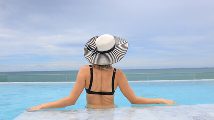 Fototapeta na wymiar Back view of the young woman in a black bikini with a straw hat on the sun-tanned slim, shapely body with her arms spread to the side, relaxing in the swimming pool on the rooftop of the hotel, enjoyi