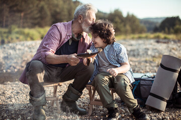 Grandfather and his grandson cooking fish they caught in a creek while out camping in the forest