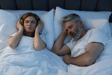 Irritated woman with headphones lying near her snoring husband in bed at home