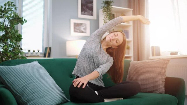 Young woman do yoga for health and wellness, meditating and stretching on sofa in beautiful living room, wearing comfortable home clothes