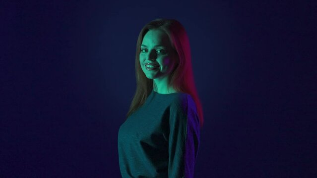 Side view of a redhaired woman in a studio against a blue background in pink and green neon light close up. The woman turns to the camera and looks straight ahead with a smile.