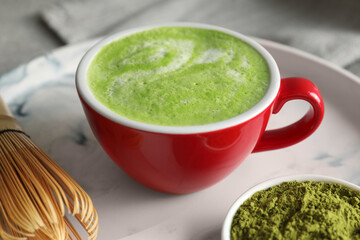 Cup of tasty matcha latte, green powder and bamboo whisk on tray, closeup