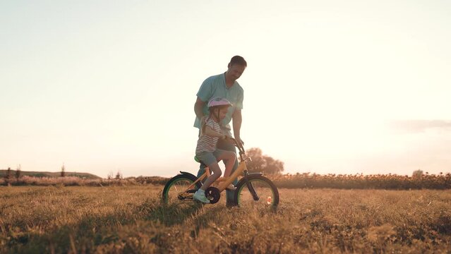 Attentive Father Supports Little Girl Learning To Ride Bicycle In Sunset Field