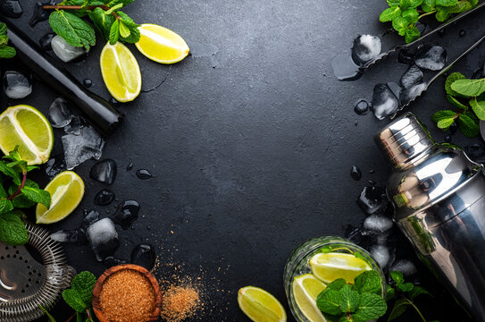 Mojito or caipirinha cocktail preparation. Mint, cane sugar, lime, ice and steel bar tools: shaker, jigger, strainer, muddler on dark stone background, top view, copy space