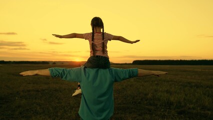 Little daughter sits on father shoulders playing plane in sunset field