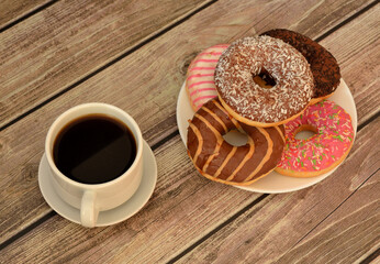 A cup of coffee on a saucer and a plate with several fresh donuts in multi-colored glaze on a light wooden table.