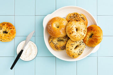 Freshly baked bagels with various toppings on a white platter. Blue tile background. Top view.