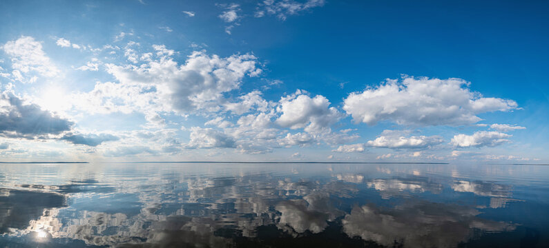 Panorama of the calm mirrored Kama River in Russia, Perm Krai, blue sky with clouds and the sun reflected in the water.