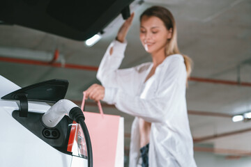Young woman travel with EV electric car to shopping center parking lot charging in downtown city...