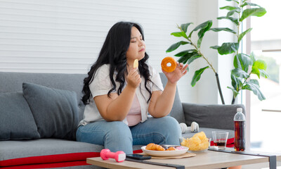 Asian young overweight oversized fat chubby plump unhealthy female teenager in casual outfit sitting thinking choosing between junk fastfood cola soft drink and dumbbell for exercise in living room - obrazy, fototapety, plakaty