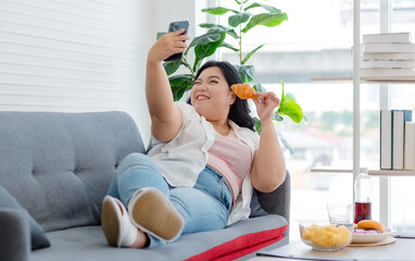 Asian young cheerful overweight oversized fat chubby plump unhealthy female teenager in casual outfit laying lying down smiling on cozy sofa holding croissant taking selfie photo via smartphone
