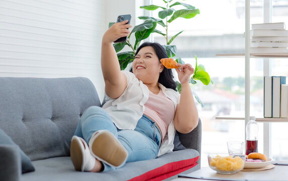 Asian Young Cheerful Overweight Oversized Fat Chubby Plump Unhealthy Female Teenager In Casual Outfit Laying Lying Down Smiling On Cozy Sofa Holding Croissant Taking Selfie Photo Via Smartphone